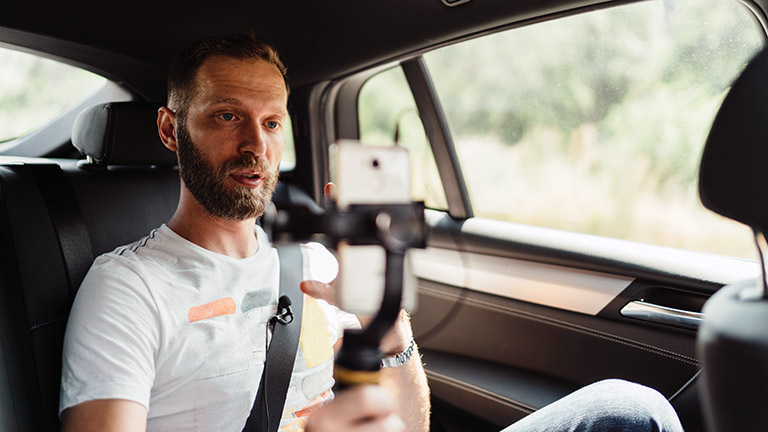 Eine Person sitzt auf der Rückbank in einem Auto und filmt sich mit dem Smartphone über ein Gimbal selbst.