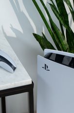 Die PlayStation 5 steht vor einer Pflanze. Daneben liegt der DualSense-Controller erhöht auf einem Tisch.