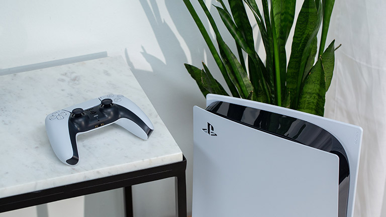 Die PlayStation 5 steht vor einer Pflanze. Daneben liegt der DualSense-Controller erh&ouml;ht auf einem Tisch.