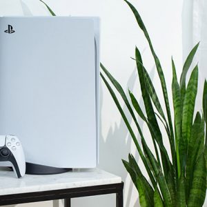 Die PlayStation 5 steht dekorativ auf einem Marmortisch neben einer Zimmerpflanze. An die Konsole angelehnt steht der neue DualSense-Controller.