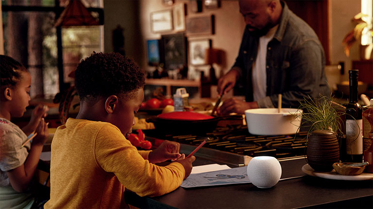 Zwei Kinder und eine Person sind in der Küche. Auf dem Tisch steht ein HomePod mini in Weiß.