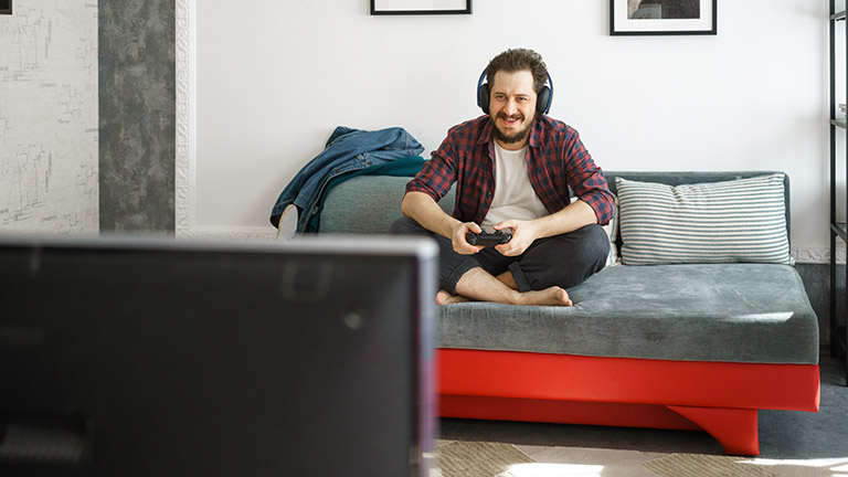 Eine Person sitzt mit Kopfhörern auf dem Sofa und spielt am Fernseher mit einer Konsole.