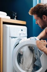Ein Mann kniet vor einer geöffneten Waschmaschine und steckt Wäsche hinein.