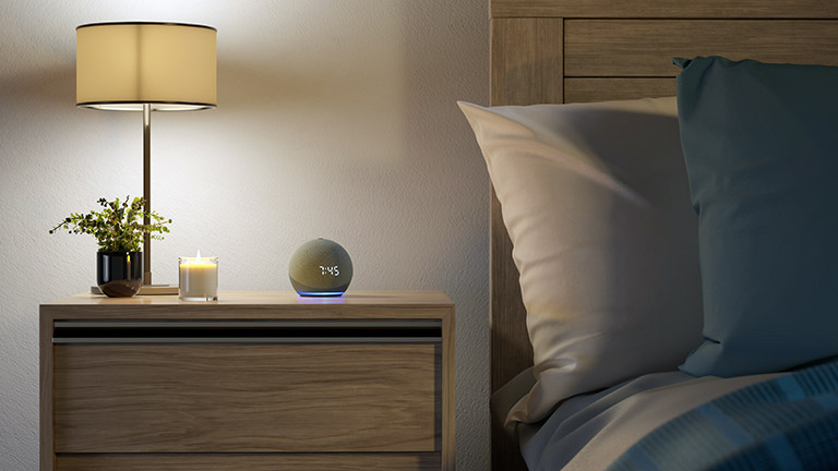 Ein Echo Dot mit Uhr-Anzeige und leuchtendem Ring neben dem Bett.