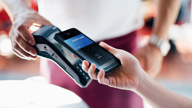 Eine Person hält ihr Smartphone an ein Kartenlesegerät und bezahlt mit einer Kreditkarte.