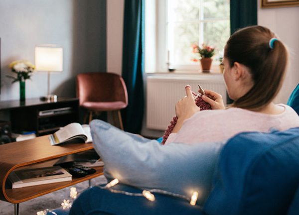 Eine Person sitzt auf dem Sofa und strickt. Nebenbei sieht sie fern.