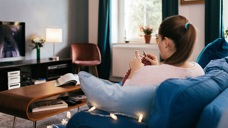 Eine Person sitzt auf dem Sofa und strickt. Nebenbei sieht sie fern.