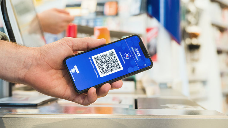 Eine Person hält ein Smartphone mit QR-Code von Payback Pay in Richtung einer Kasse.