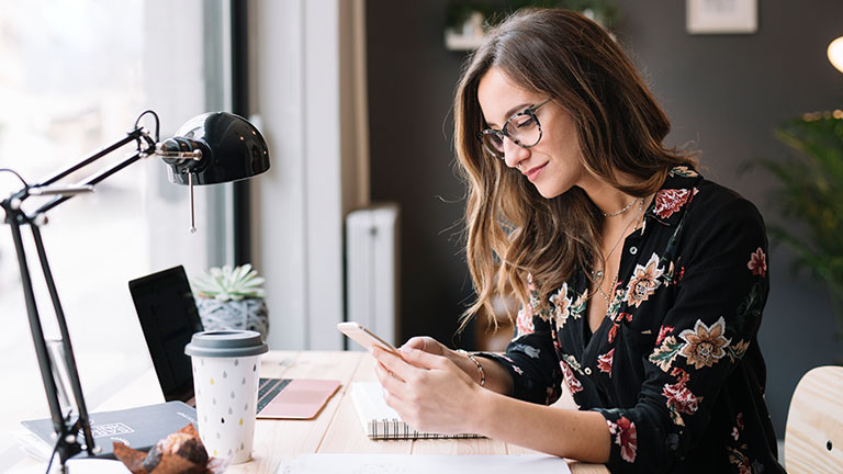 Eine Person sitzt an einem Schreibtisch und hält ein Smartphone in der Hand.