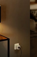 Ein Apple HomePod Mini auf einem Tisch. Daneben steht eine Lampe, die über eine Eve Energy mit dem Strom verbunden ist.