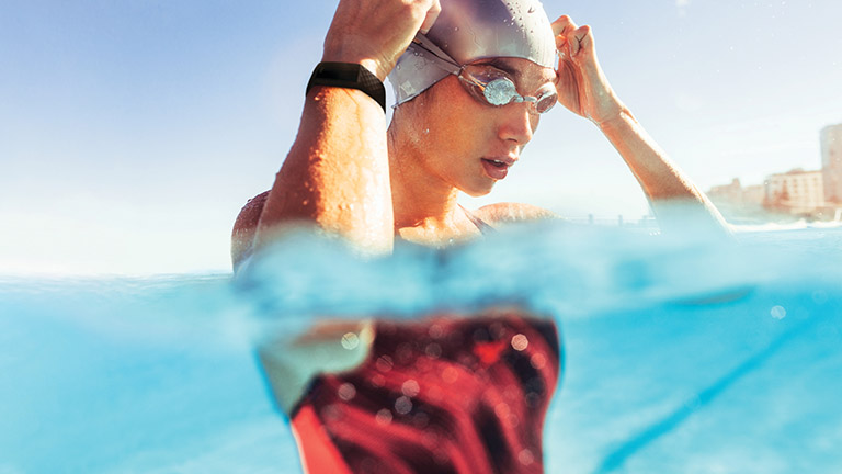 Eine Schwimmerin im Wasser setzt sich ihre Schwimmbrille auf. Am Handgelenk trägt sie das wasserabweisende Fitness-Armband Fitbit Charge 4.