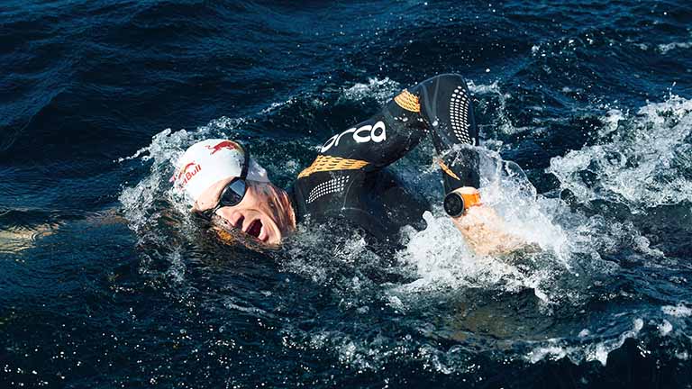 Ein Leistungsschwimmer beim Kraulschwimmen. An seiner linken Hand, die gerade aus dem Wasser schnellt, trägt er die Polar Vantage V mit orangenem Armband.