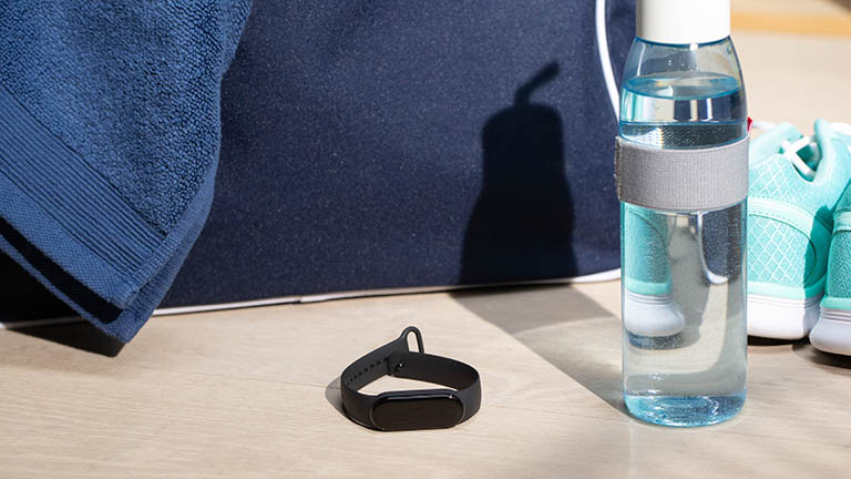 Das Fitness-Armband Mi Smart Band 5 von Xiaomi liegt vor einer großen, blauen Sporttasche. Daneben befinden sich eine Wasserflasche und ein Paar Sportschuhe.