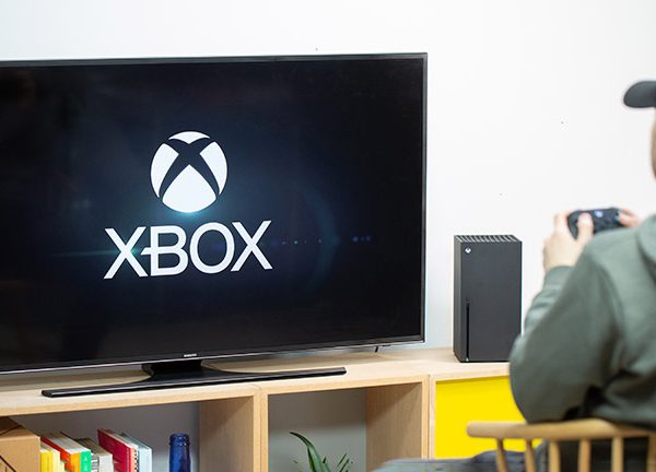 Eine Person sitzt vor einer Xbox Series X. Auf dem Fernseher ist der Startbildschirm zu sehen.