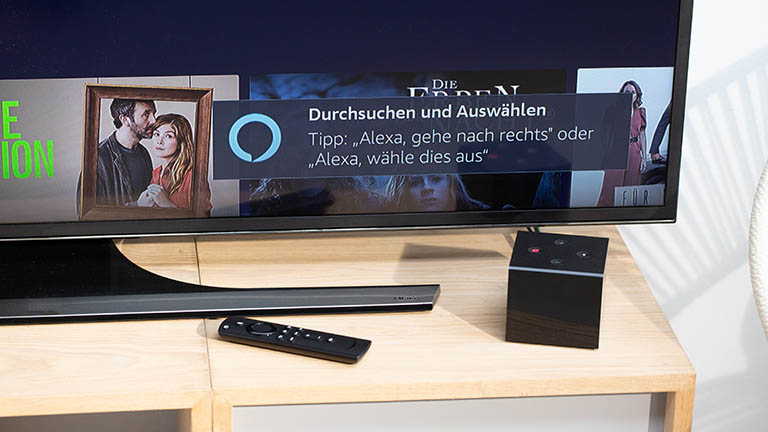 Ein TV-Gerät, auf dem ein Tipp für die Steuerung per Alexa angezeigt ist. Vor dem Fernseher steht der Fire TV Cube.