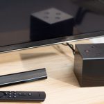 Der Amazon Fire TV Cube steht vor einem Fernseher. Daneben ist die Fernbedienung zu sehen.