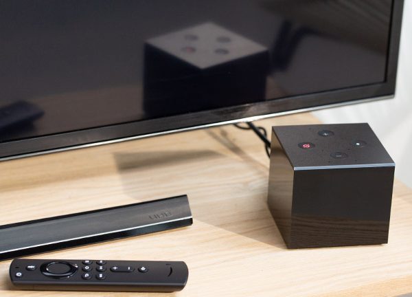Der Amazon Fire TV Cube steht vor einem Fernseher. Daneben ist die Fernbedienung zu sehen.
