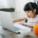 Ein junges M&auml;dchen sitzt an einem K&uuml;chentisch vor einem aufgeklappten Laptop. Neben ihr befinden sich ein paar Stifte und ein volles Glas Saft.
