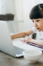 Ein junges Mädchen sitzt an einem Küchentisch vor einem aufgeklappten Laptop. Neben ihr befinden sich ein paar Stifte und ein volles Glas Saft.