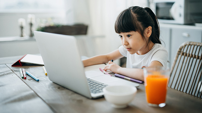 Ein junges Mädchen sitzt an einem Küchentisch vor einem aufgeklappten Laptop. Neben ihr befinden sich ein paar Stifte und ein volles Glas Saft.