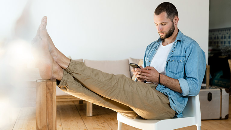 Eine Person sitzt auf einem Stuhl mit dem Smartphone in der Hand und legt die Füße hoch.