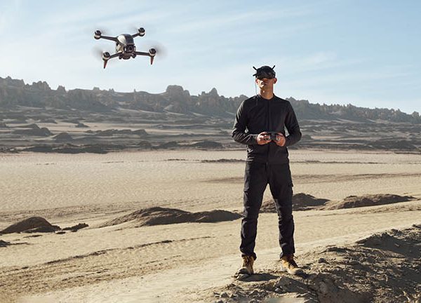 Ein Mann steht in sandiger Umgebung. Er h&auml;lt einen Drohnencontroller in der Hand und tr&auml;gt die FPV-Brille von DJI. Vor ihm fliegt die DJI FPV.
