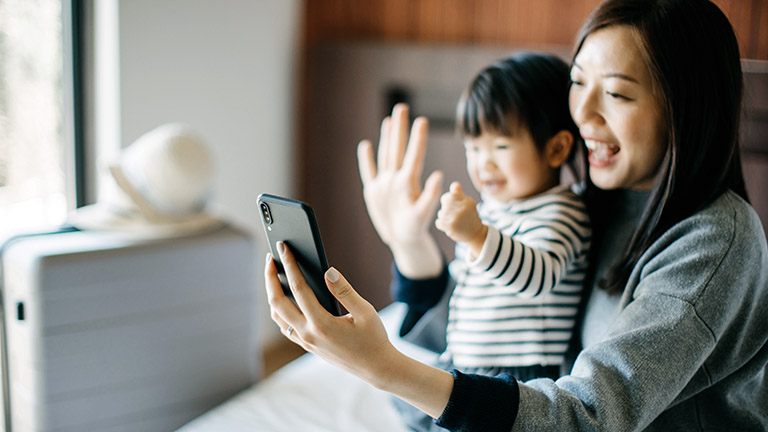 Eine Person und ihr Kind schauen in die Kamera eines Smartphones und winken dabei.
