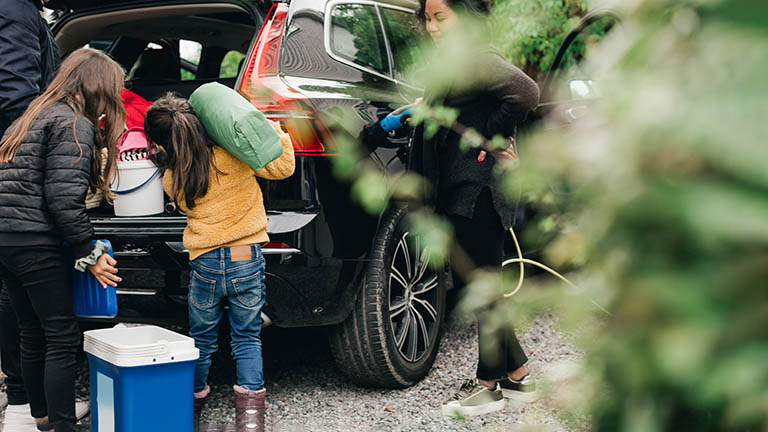 Eine Familie belädt den Kofferraum eines Autos für einen Ausflug, auf dem Boden davor steht eine thermoelektrische Kühlbox.