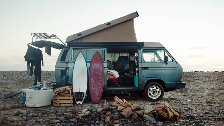 An einem Strand steht ein Camping-Van, die Seitentür ist geöffnet, eine mobile Kühlbox steht daneben.