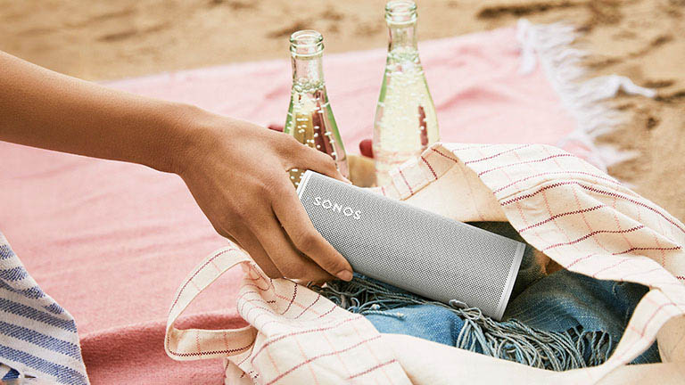 Eine Hand zieht den Sonos Roam aus einer Handtasche. Die Person sitzt dabei auf einer Picknickdecke. Im Hintergrund sind zwei Getränkeflaschen zu erkennen.