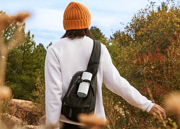 Ein Mann streift durch ein Feld. An seinem Rucksack h&auml;ngt der Bluetooth-Lautsprecher Sonos Roam in wei&szlig;.