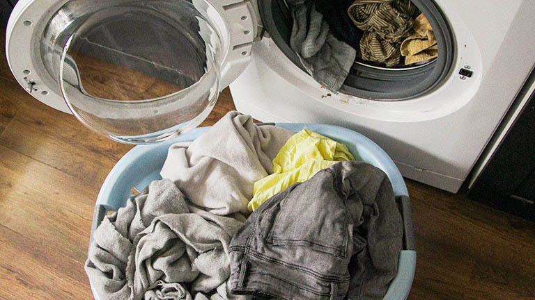 Eine offene Waschmaschine, in der noch etwas Wäsche steckt. Davor steht ein Korb mit weiterer Wäsche.
