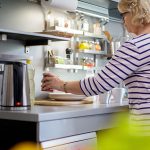 Eine Person steht in der K&uuml;che, auf der Arbeitsplatte steht ein eingeschalteter Wasserkocher.