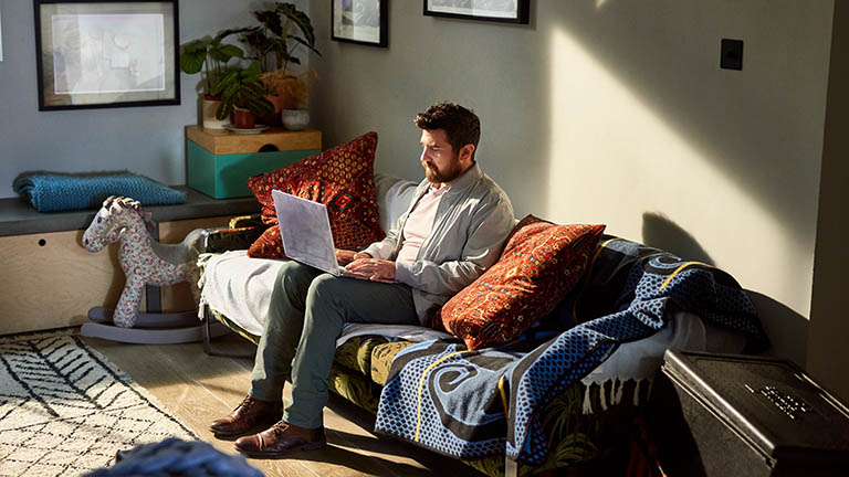 Ein Mann sitzt auf einem Sofa, auf seinen Knien balanciert er ein aufgeklapptes Notebook, seine Hände ruhen auf der Tastatur.