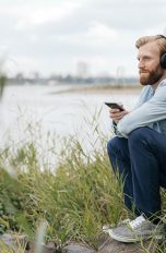 Eine Person sitzt vor einem Fluss auf einem Stein und hört über das Smartphone mit Kopfhörern Musik.