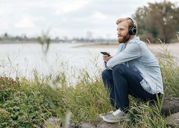Eine Person sitzt vor einem Fluss auf einem Stein und hört über das Smartphone mit Kopfhörern Musik.