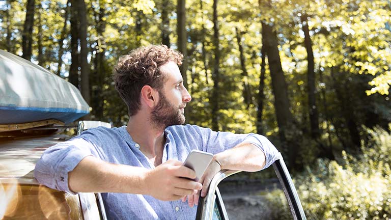 Eine Person steht in der Autotür, hält ihr Smartphone in der Hand und schaut in die Natur.