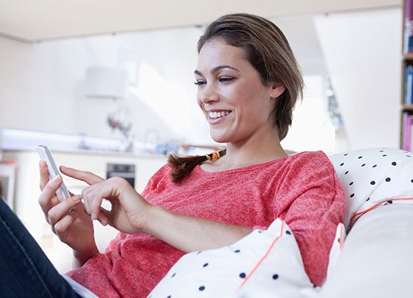 Eine Person sitzt entspannt auf einem Sofa und tippt l&auml;chelnd auf ein Smartphone.