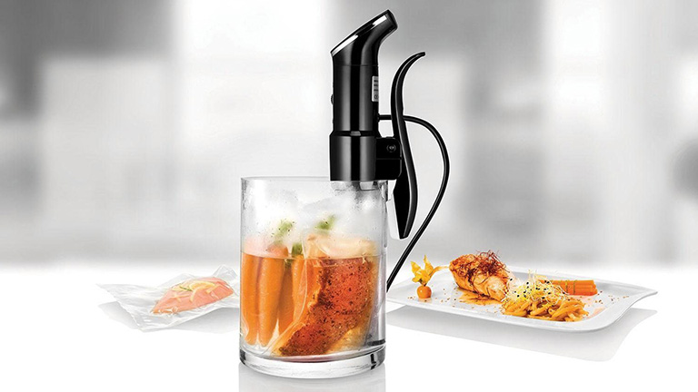Unold Dampfgarer Sous-Vide-Stick Time 58915 Der Unold Sous-Vide-Stick Time 58915 steckt in einem Wasserbehälter als Glas, in dem sich ein Garbeutel mit Möhren und Lachs befindet, im Hintergrund befindet sich ein angerichteter Teller.