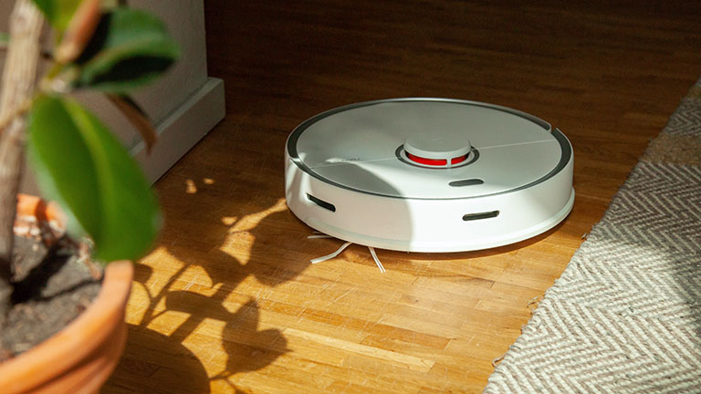 Ein Roborock S5 Max f&auml;hrt &uuml;ber einen Holzboden auf eine Topfpflanze zu.