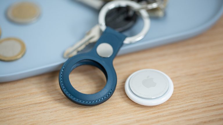 Ein Apple AirTag liegt neben einem Schlüsselanhänger. Im Hintergrund sind ein paar Euro-Münzen für den Größenvergleich zu sehen.)