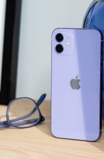 Das iPhone 12 in der neuen Farbe Violett steht auf einem Holztisch. Daneben liegt eine farblich passende Brille.