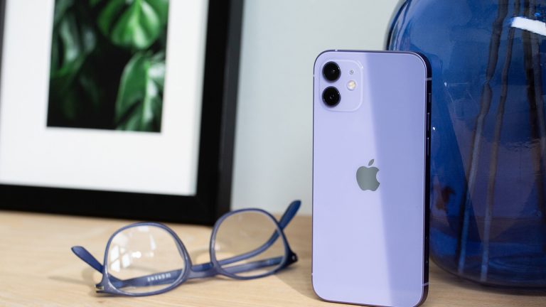 Das iPhone 12 in der neuen Farbe Violett steht auf einem Holztisch. Daneben liegt eine farblich passende Brille.