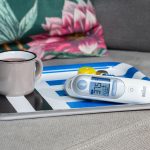 Auf einem Tablett steht eine Emailletasse mit Tee, daneben liegt ein Infrarotfieberthermometer.