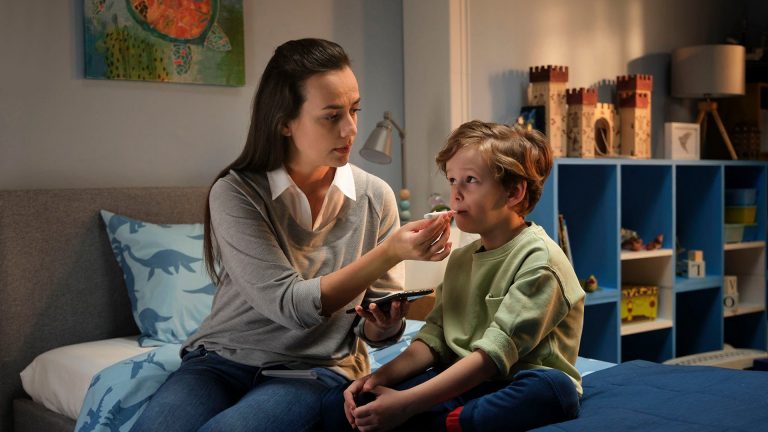 Eine Mutter sitzt mit ihrem Sohn auf der Bettkannte und hält das Fieberthermometer, das in seinem Mund steckt.