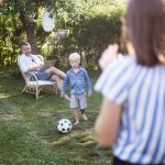 Eine Person sitzt auf einem Stuhl im Garten, w&auml;hrend ihr Sohn Anlauf nimmt, um einen Ball zu schie&szlig;en, eine weitere Person steht im Vordergrund und schaut zu.