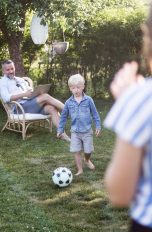 Eine Person sitzt auf einem Stuhl im Garten, während ihr Sohn Anlauf nimmt, um einen Ball zu schießen, eine weitere Person steht im Vordergrund und schaut zu.