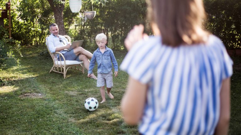 Eine Person sitzt auf einem Stuhl im Garten, w&auml;hrend ihr Sohn Anlauf nimmt, um einen Ball zu schie&szlig;en, eine weitere Person steht im Vordergrund und schaut zu.