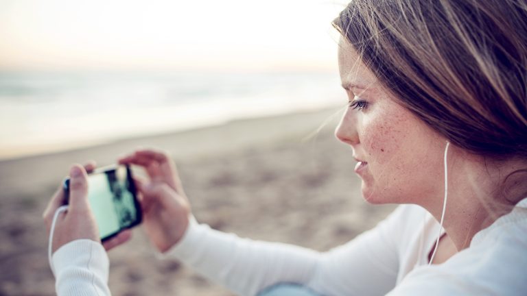 Eine Person sitzt am Meer und schaut auf ihrem Smartphone einen Videostream.