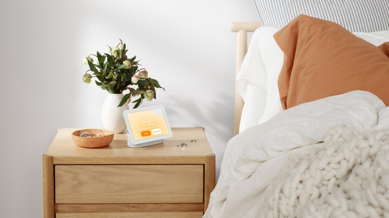 Der Google Nest Hub steht auf einem Nachttisch neben einem Bett.